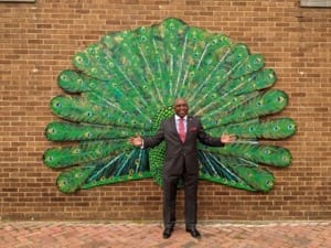 A man in a suit poses in front of the Portsmouth Public Art Wings entitled "Proud Peacock"