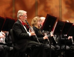 Two people within a large band play clarinets while reading music