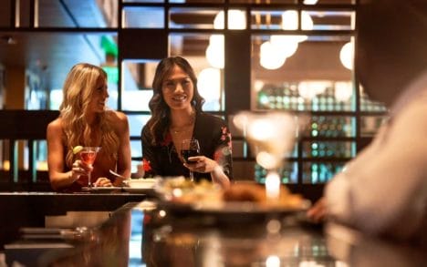 Two women sip drinks and talk to a man at a bar.