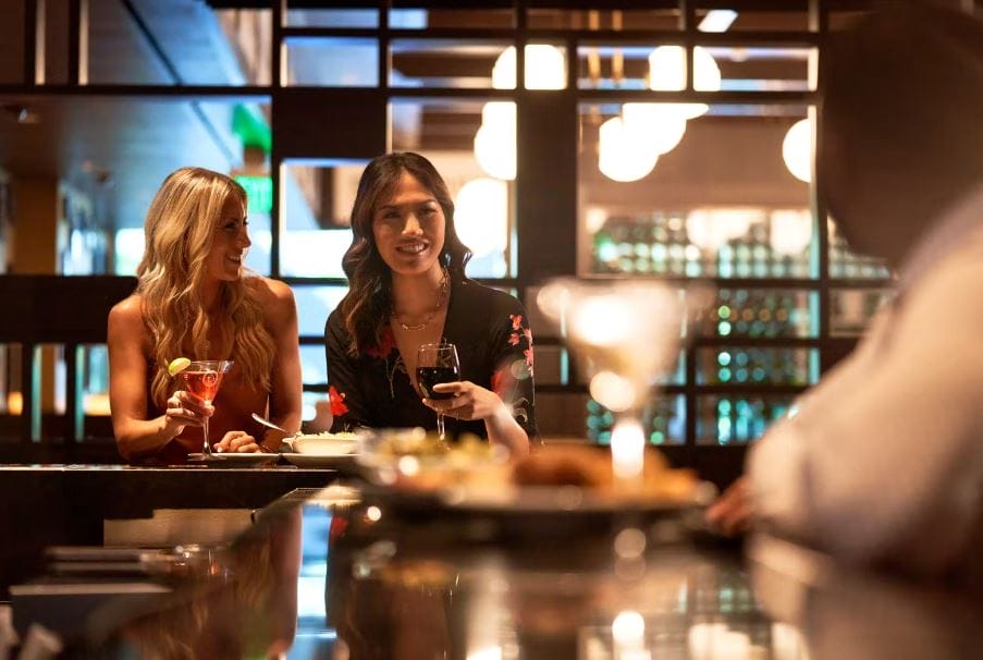 Two women sip drinks and talk to a man at a bar.