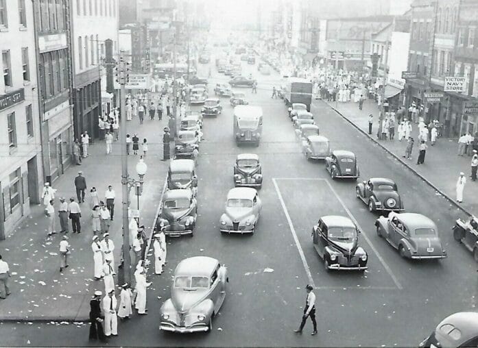 Black and White image from the 1940's in Olde Towne Portsmouth, VA. Photo shows old cars dring around the street and civilians and sailors walking the sidewalks.