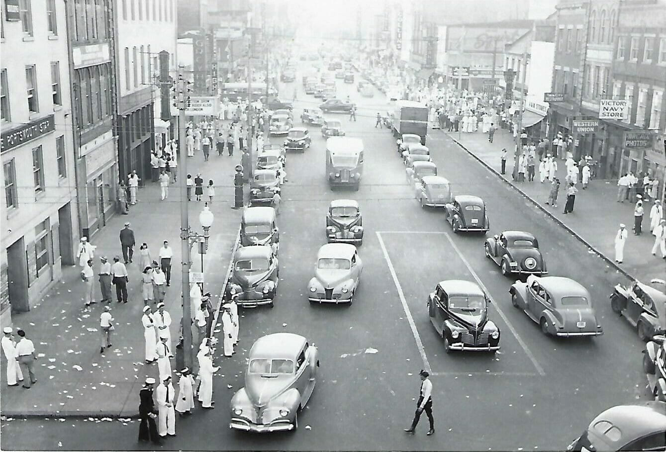 Black and White image from the 1940's in Olde Towne Portsmouth, VA. Photo shows old cars dring around the street and civilians and sailors walking the sidewalks.