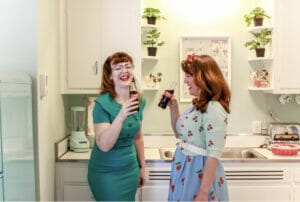 two women dressed in mid-century dresses laughing and drinking a Coke in a 50s era kitchen