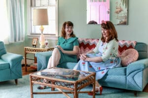 two women sitting on 50s era couch dressed in 50s era attire.